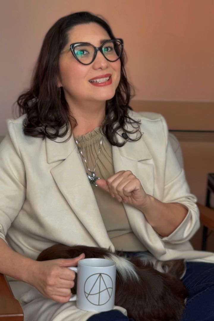 Dra. Angela Mori sorrindo, sentada numa poltrona, com uma caneca da Anima Integri e seu cachorro no colo