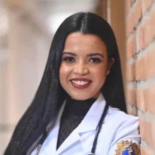 Dra. Larissa Guerra sorrindo, de jaleco branco e estetoscópio, encostada numa parede de tijolos